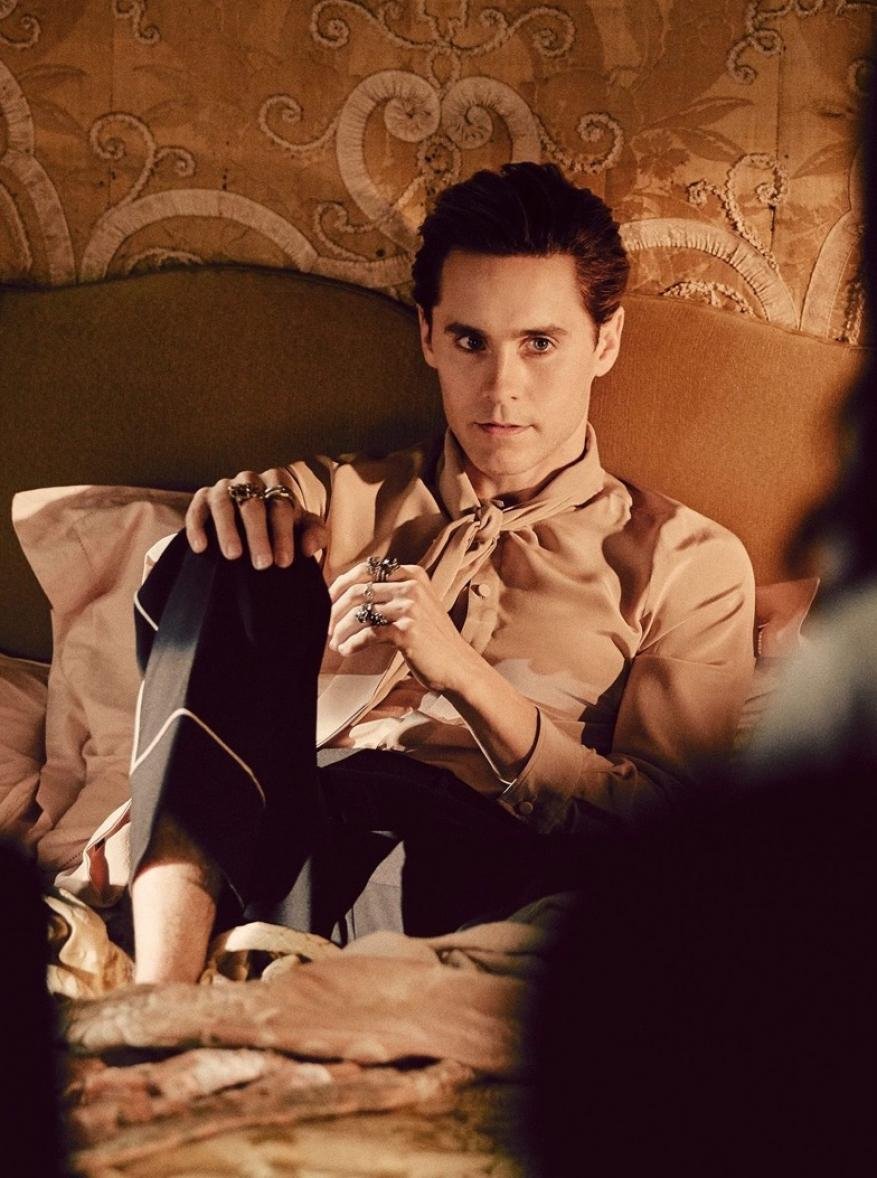 Indoor portrait of adult male wearing rings, silk shirt, and dress pants lying on a bed against an ornate patterned wallpaper.