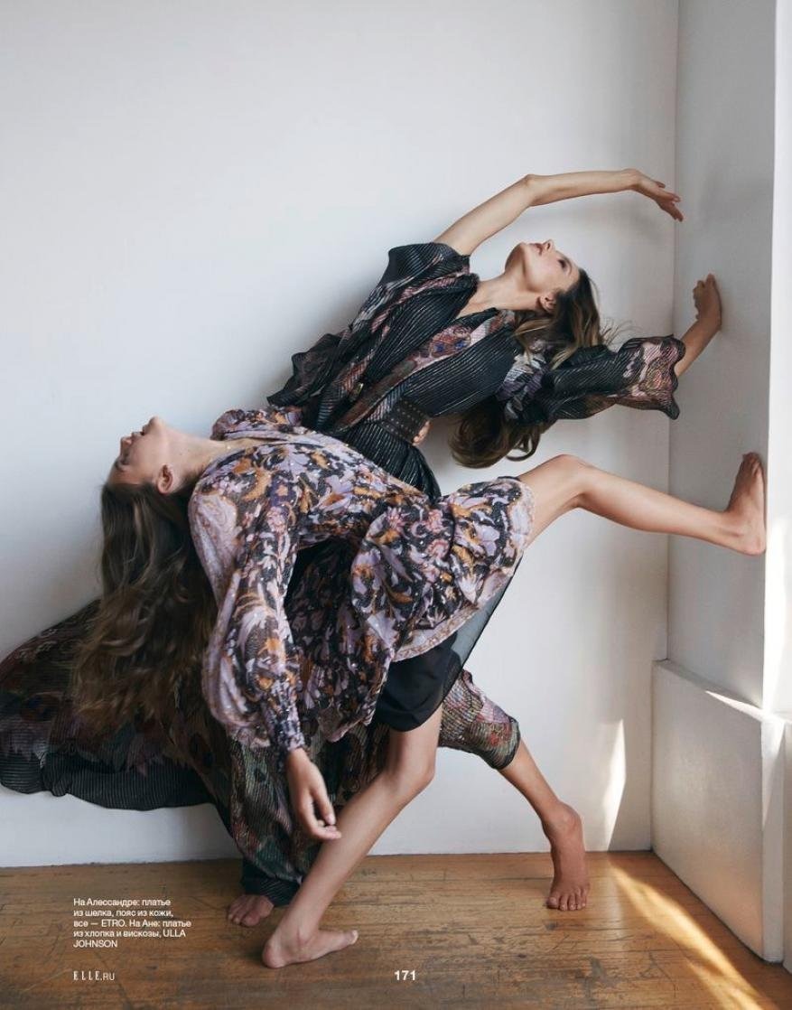 Two women strike a pose in flowing floral dresses against a minimalist wall, showcasing fashion in a studio setting.