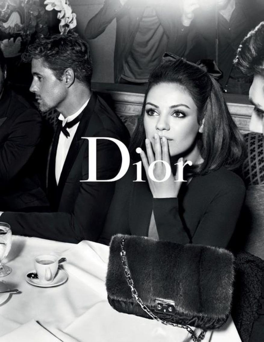 Black and white fashion shot of Mila Kunis sitting at a table with other people in suits in the background, holding her hand up to her face.