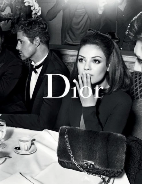 Black and white fashion shot of Mila Kunis sitting at a table with other people in suits in the background, holding her hand up to her face.
