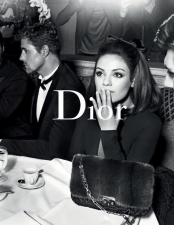 Black and white fashion shot of Mila Kunis sitting at a table with other people in suits in the background, holding her hand up to her face.