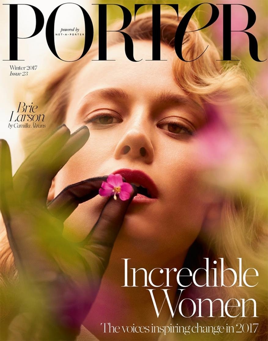 Close-up magazine cover shot of Brie Larson with finger to lip, holding flower, against blurred purple-green foliage backdrop.