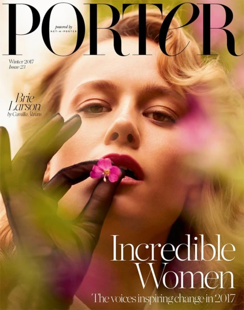 Close-up magazine cover shot of Brie Larson with finger to lip, holding flower, against blurred purple-green foliage backdrop.