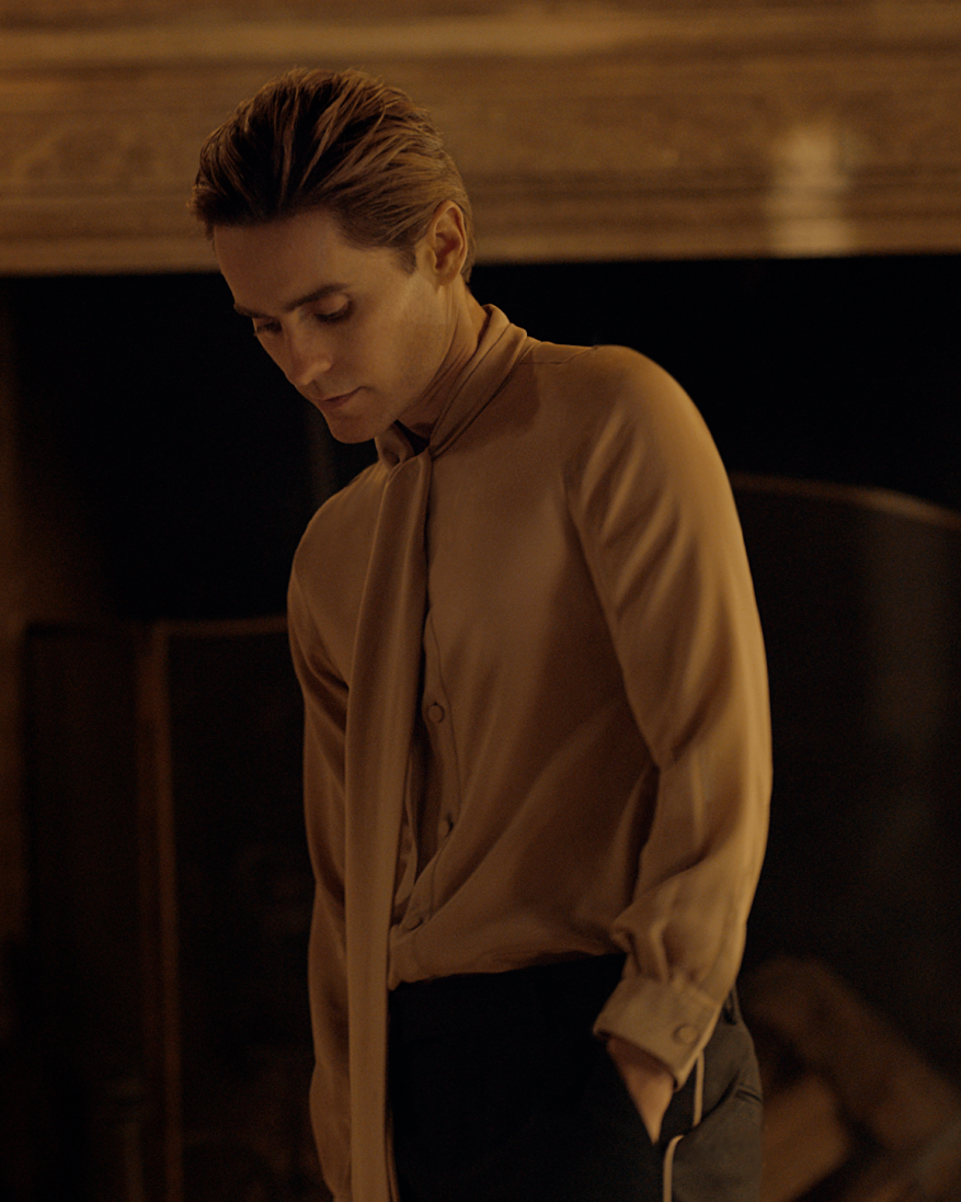 Headshot of a blonde man with slicked-back hair wearing a light brown blouse with a neck tie. He is looking down and posing.