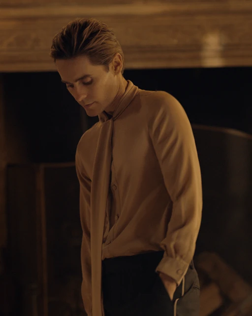 Adult male with styled blond hair in a beige, tie-neck shirt and black pants poses indoors in a dimly lit setting with dark, neutral tones.