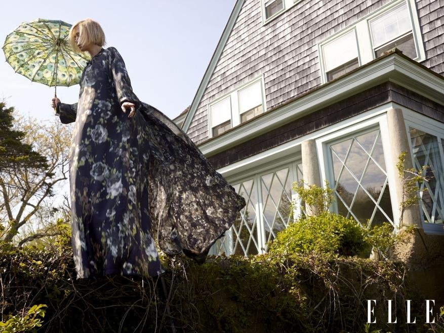 A blonde woman in a floral dress is holding an umbrella outside a house on a sunny day, showcasing fashion and outdoor style with elegance.