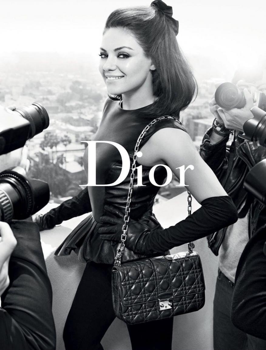 Fashion shot of Mila Kunis in a Dior campaign, posing with a black quilted bag, surrounded by photographers in a studio setting.