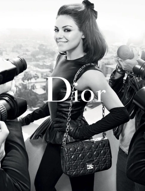 Fashion shot of Mila Kunis in a Dior campaign, posing with a black quilted bag, surrounded by photographers in a studio setting.