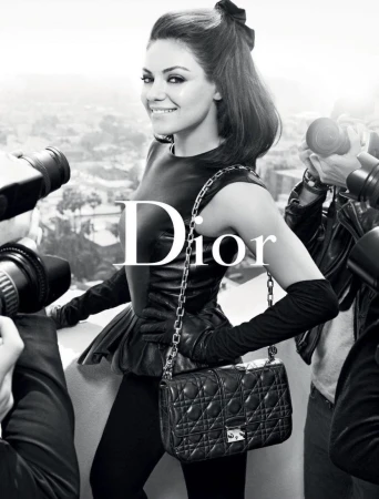 Fashion shot of Mila Kunis in a Dior campaign, posing with a black quilted bag, surrounded by photographers in a studio setting.