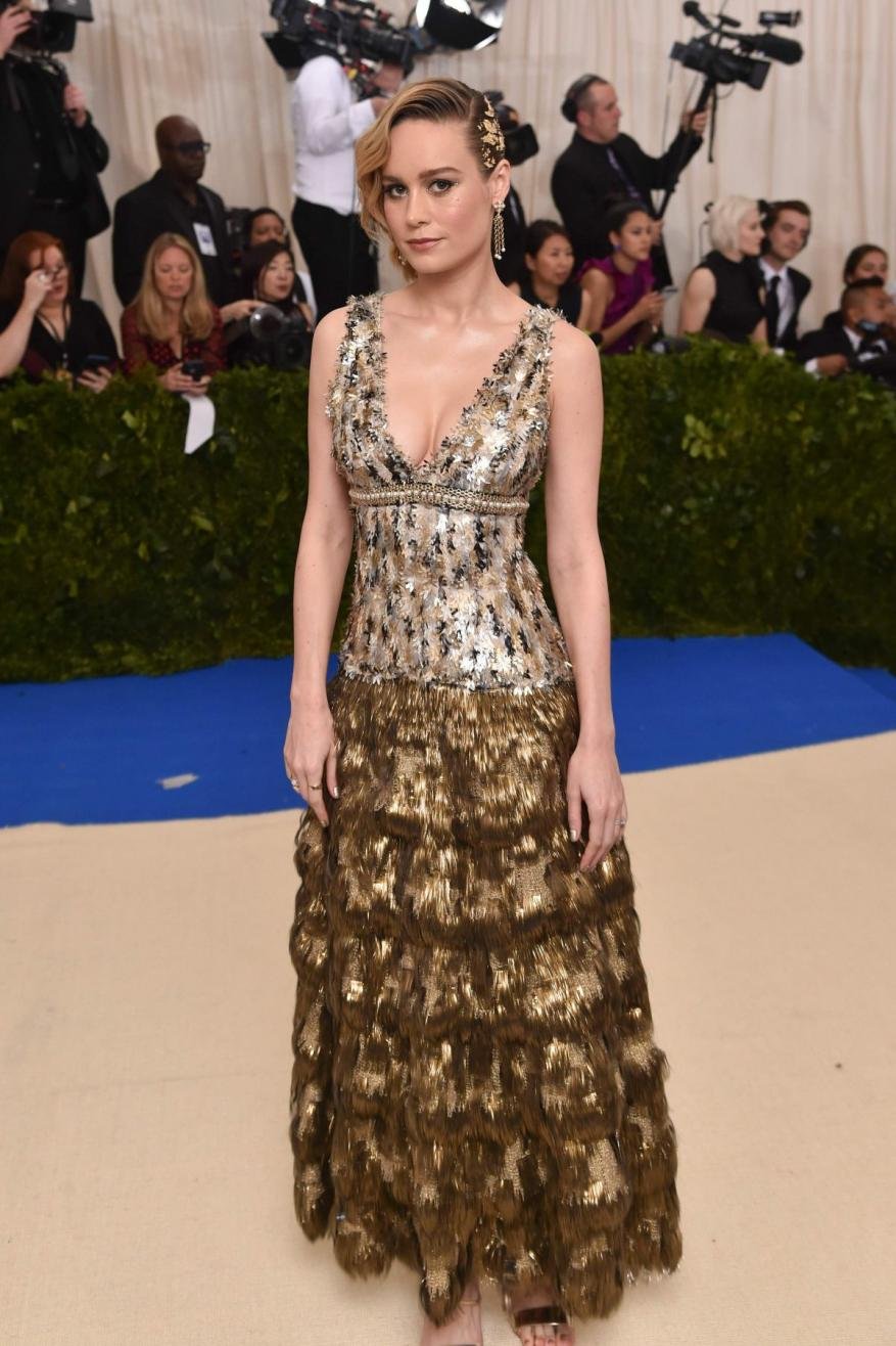 Brie Larson poses on the red carpet in a gold and silver floor-length gown, with media and photographers in the background.