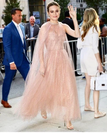 Brie Larson on the red carpet in a peach gown, smiling, and waving, surrounded by onlookers and barriers outside a building.