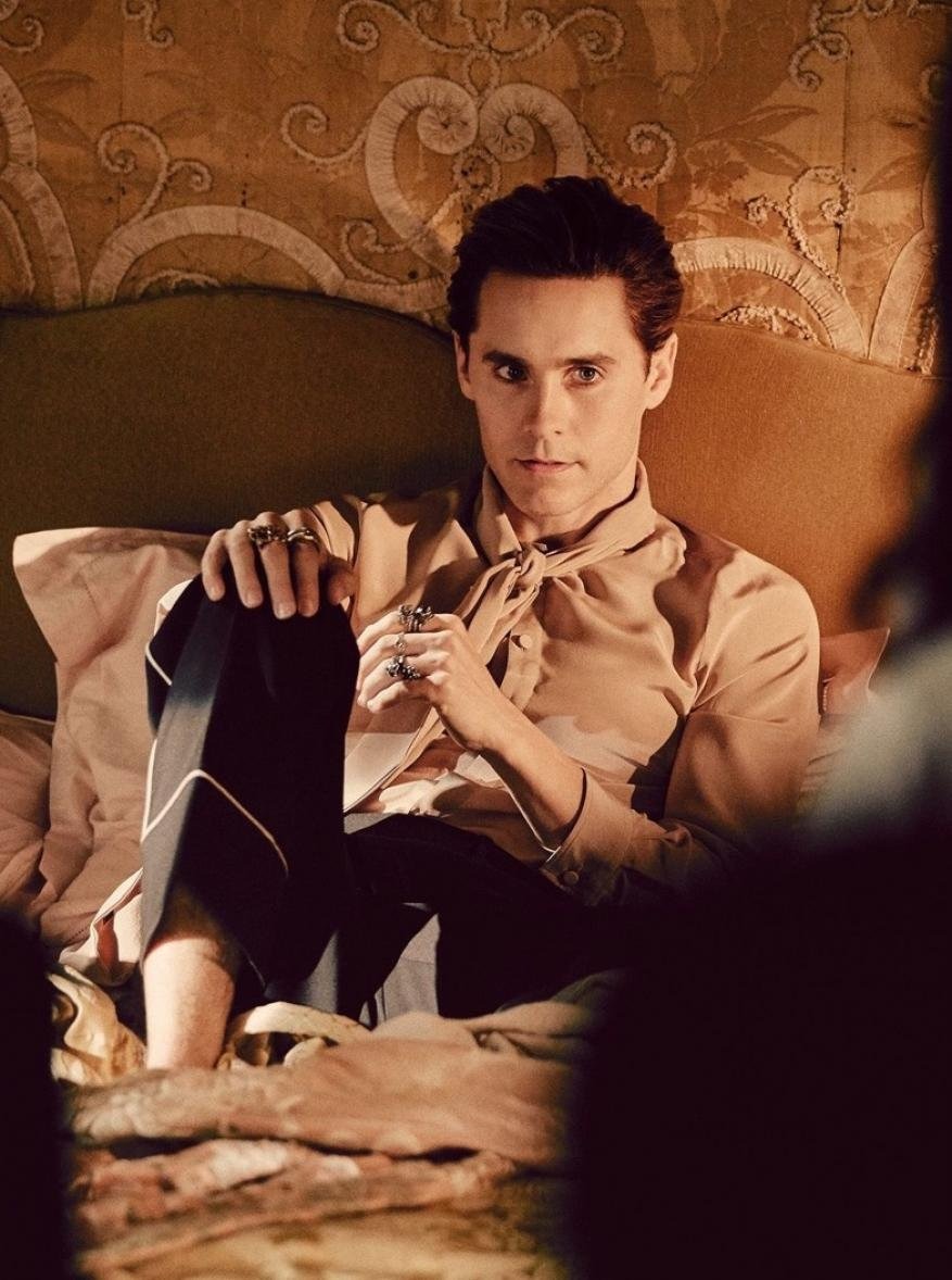 Adult man wearing jewelry and tan button-down shirt with a tied collar in bed, set against patterned wallpaper backdrop.
