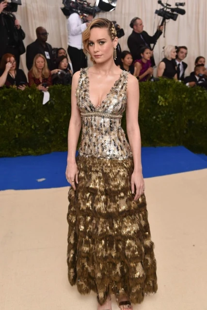 Brie Larson on the red carpet at the Met Gala, wearing a tiered gold sequin gown. The background shows a crowd with camera equipment.