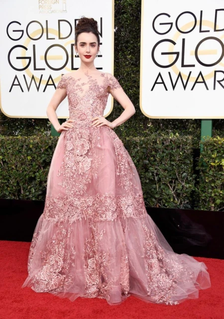 Lily Collins in a pale pink lace gown poses on the red carpet at the Golden Globes, with signage and hedge in the background.