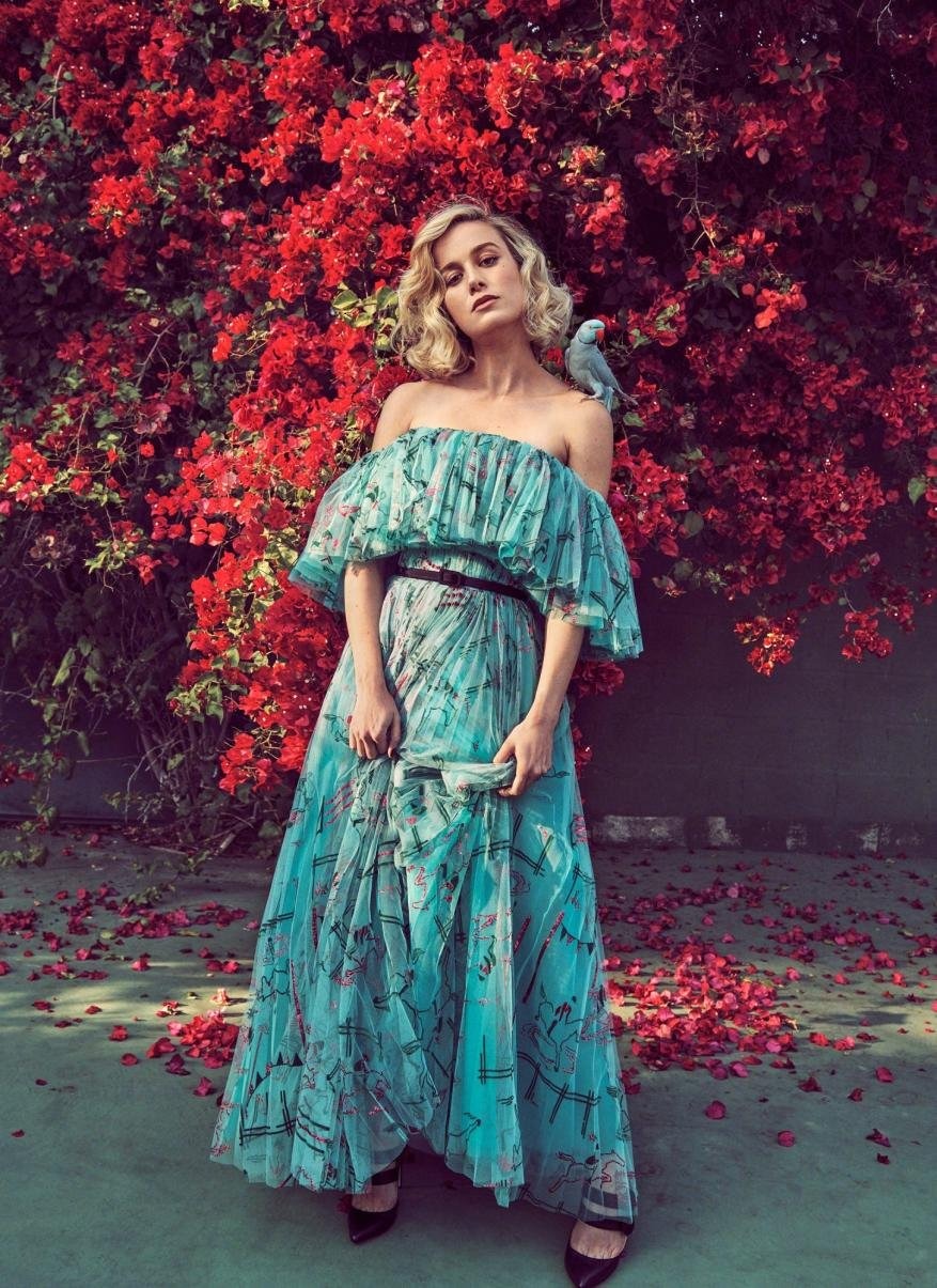 Brie Larson in a flowing turquoise gown posing outdoors with a parrot perched on her shoulder against a backdrop of vibrant red flowers.