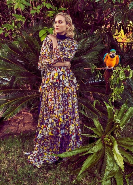 A woman in a floral dress stands next to a perched macaw parrot in a lush, tropical garden setting with ferns and leafy foliage.