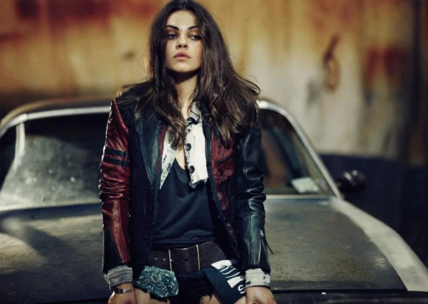 Mila Kunis poses in a black and red leather jacket in front of a vintage car with a blurred urban background, styled in a casual fashion look.