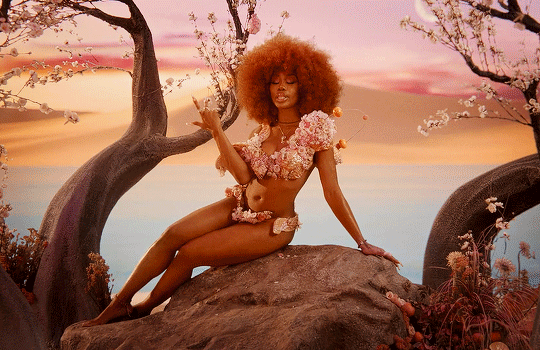 A woman with afro hairstyle poses in a floral bikini on a rock in front of a pastel sunset sky with blossoming trees.