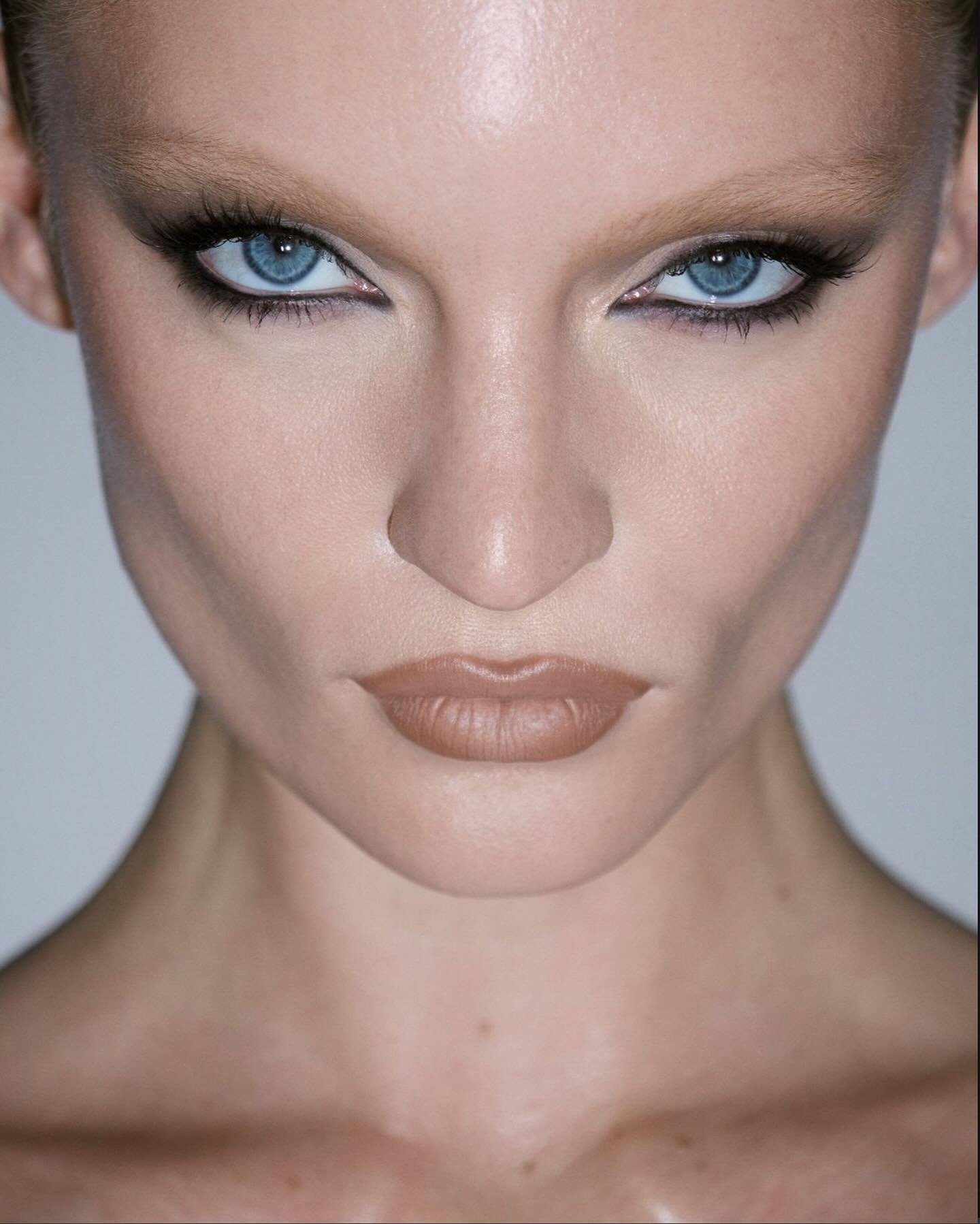 Close-up studio shot of a fair-skinned young woman with blue eyes wearing dramatic eye makeup and nude lipstick looking directly at camera.