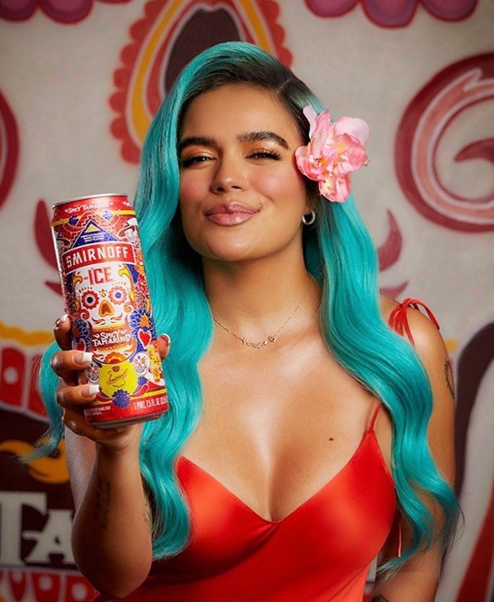 Adult female with aqua hair poses indoors in front of a floral backdrop, holding a Smirnoff Ice Tamarindo can in a medium shot.