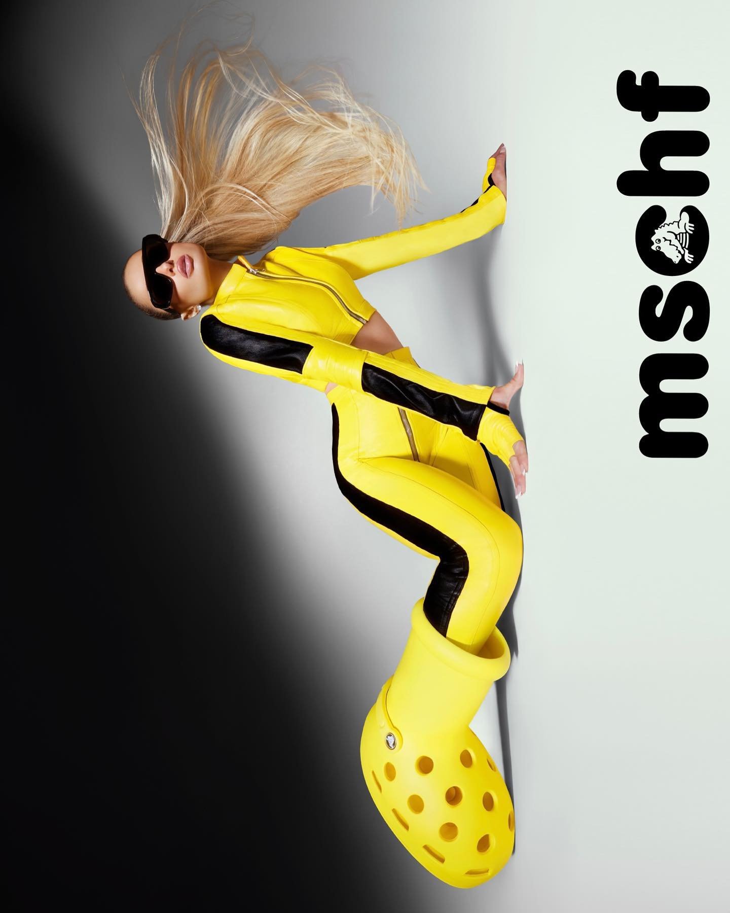Woman in yellow jumpsuit and oversized Crocs poses against a split white and black background, wearing sunglasses with flowing blonde hair.