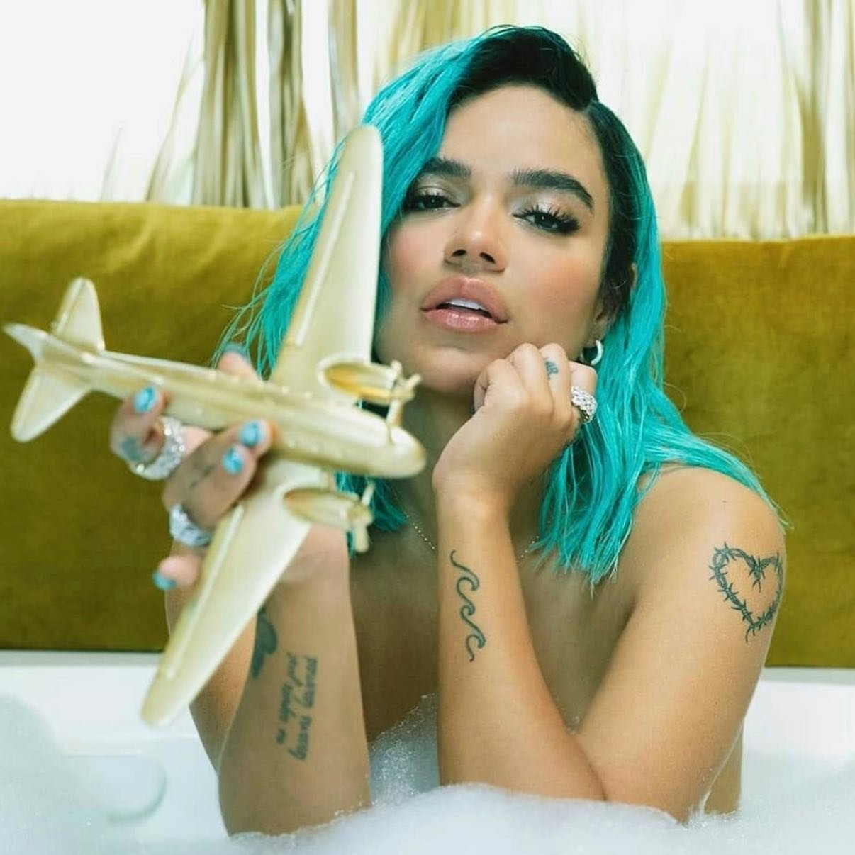 Portrait of a woman with turquoise hair in a bathtub holding a golden toy airplane, set against an olive-green cushioned headboard.