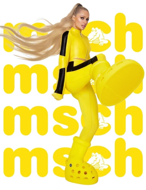 Paris Hilton poses in bright yellow outfit and oversized Crocs, a fashion-forward and playful image against a logo background.