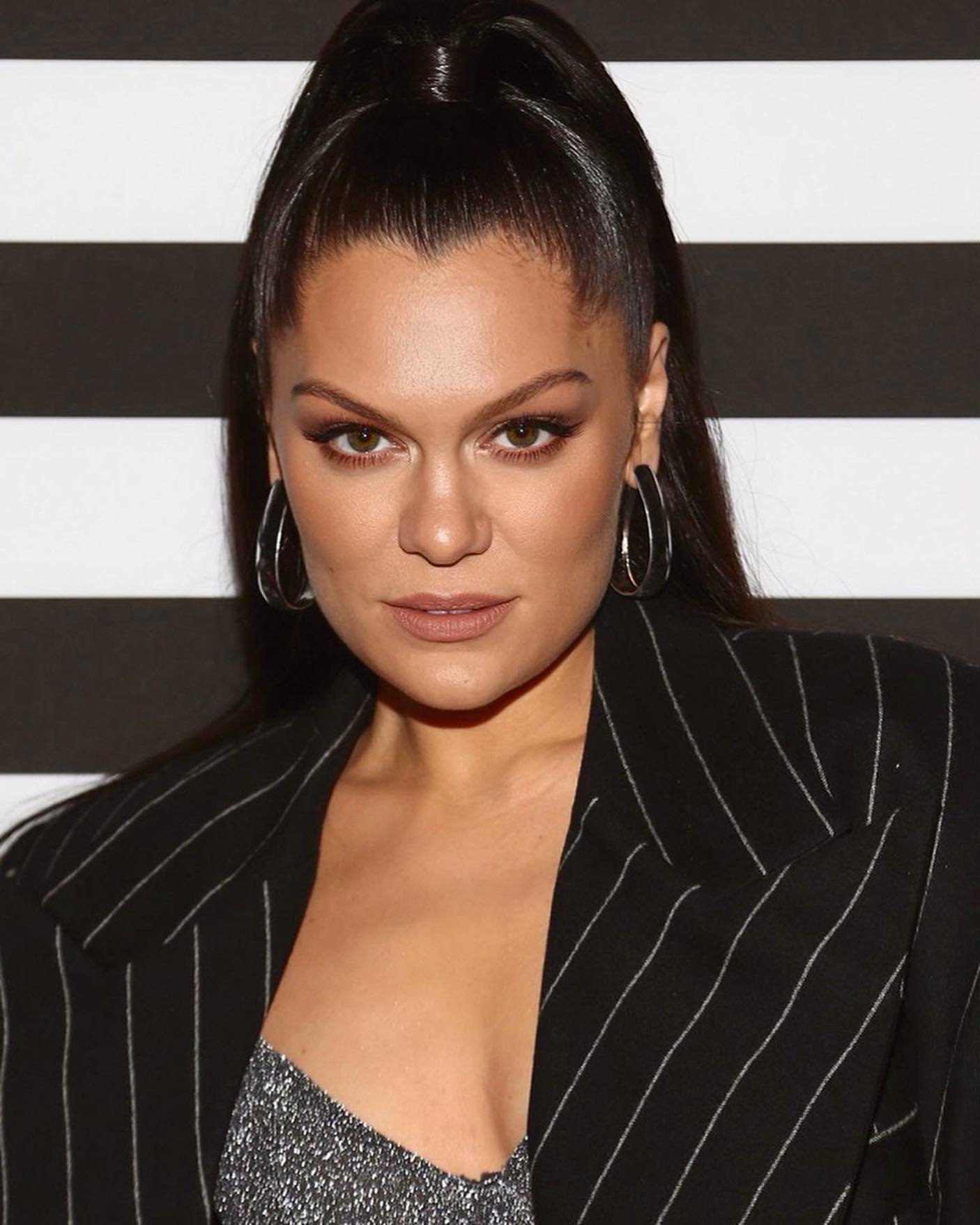 Close-up of a woman with dark hair in a high ponytail wearing hoop earrings and a black pinstripe jacket against a striped backdrop.