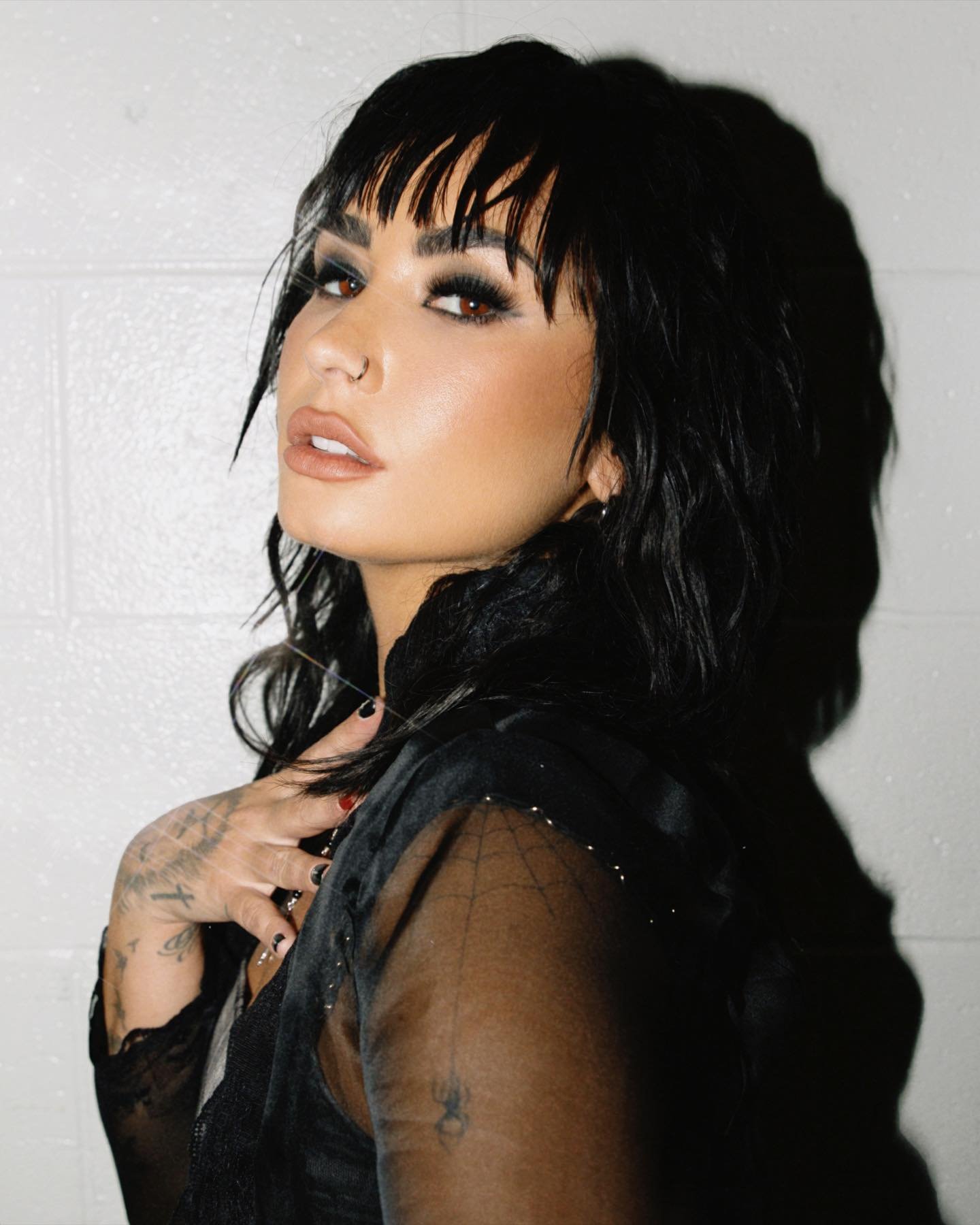 Portrait of Demi Lovato with dark hair and smokey eye makeup wearing a sheer black top against a white textured background.