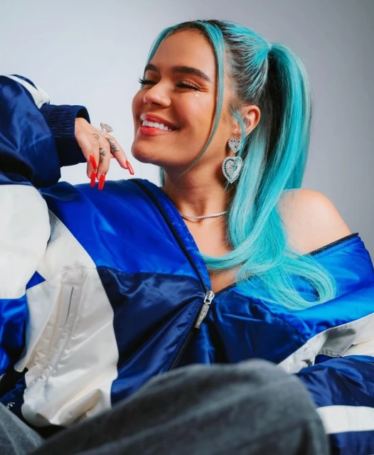 Close-up studio portrait of a smiling adult female with blue hair wearing a colorful jacket and heart-shaped earrings, sitting indoors.