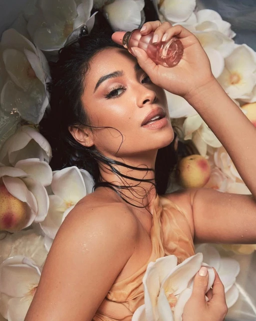 Woman with flowers in beauty shot. The model holds a cosmetic product, surrounded by white flowers, peach, and a nude colored fabric.