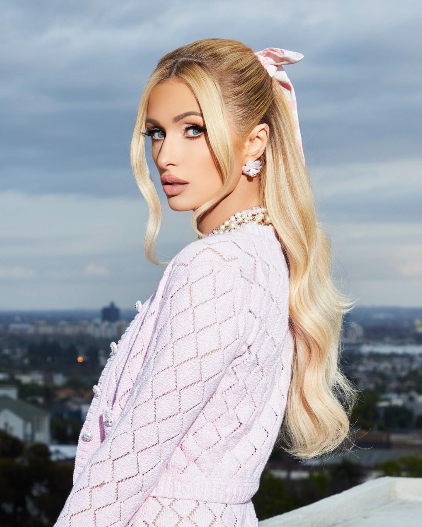 Blonde woman in lilac cardigan and skirt with pearl jewelry stands outdoors with blurred city background and pale blue sky.