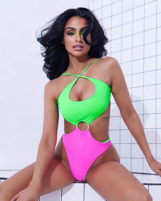 Brunette woman in a vibrant neon green and pink one-piece swimsuit poses against a tiled wall, showcasing bold makeup and an indoor studio setting.