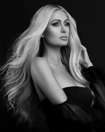 Portrait of Paris Hilton in black and white with voluminous blonde hair and dark off-the-shoulder top against a dark background.