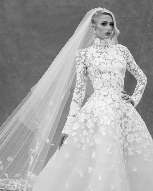 Black-and-white portrait of a woman in a lace wedding gown and veil, posing against a textured backdrop for a formal portrait.