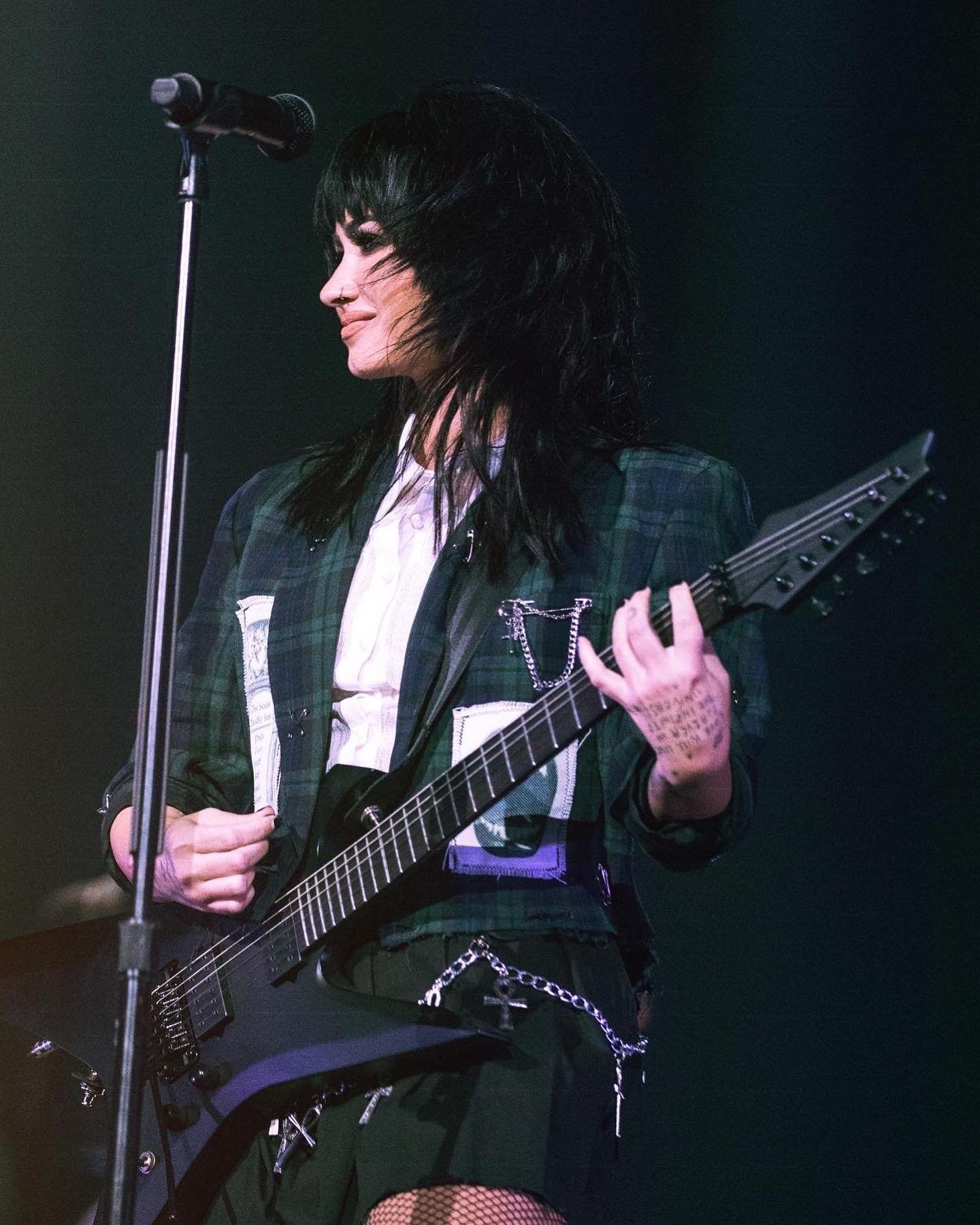 A young adult female plays an electric guitar on stage, wearing a plaid jacket and dark clothing, with a microphone stand beside her.
