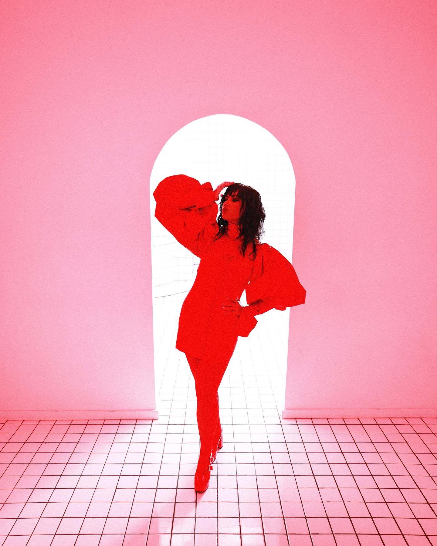 A woman poses in a vibrant red dress with puffy sleeves, set against a doorway in a pink-toned interior with tiled flooring.