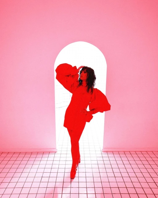A woman poses in a vibrant red dress with puffy sleeves, set against a doorway in a pink-toned interior with tiled flooring.