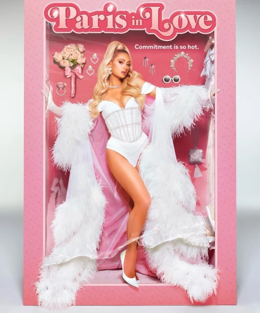 Paris Hilton appears as a doll in a pink toy box wearing a white outfit, a white robe with feathered trim, and matching heels.