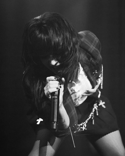 Monochrome concert shot features an adult female vocalist holding a microphone, dressed in patterned blazer with a dark background.