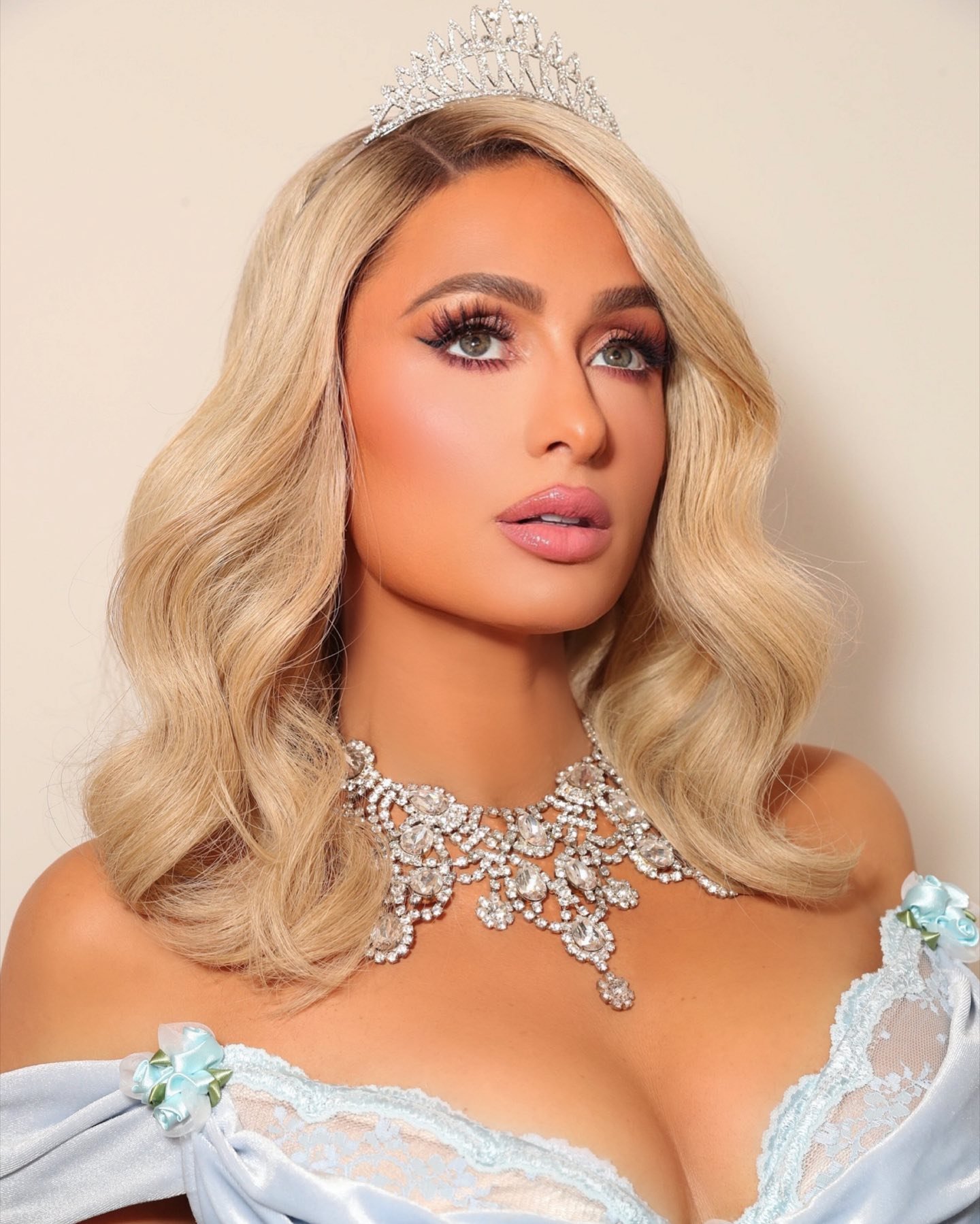 Close-up portrait of Paris Hilton in an off-the-shoulder dress, diamond tiara, and elaborate diamond necklace against a neutral backdrop.
