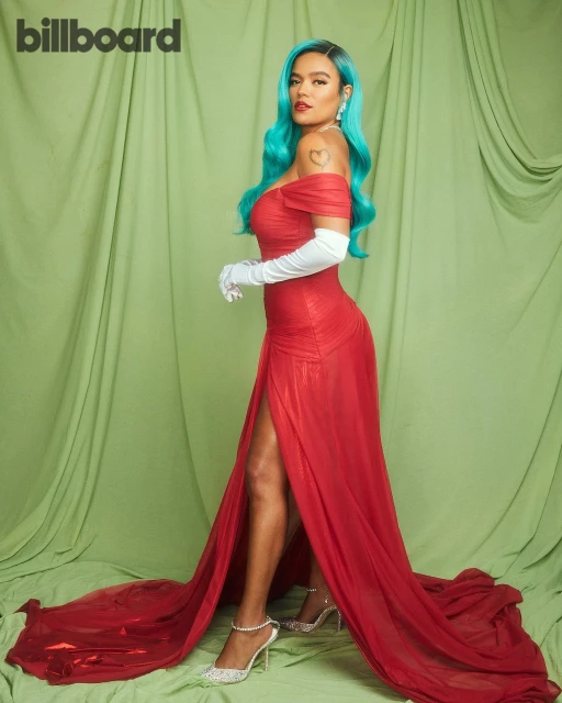 Karol G in a full-length pose with blue hair, wearing a red dress and white gloves in front of a light green background.
