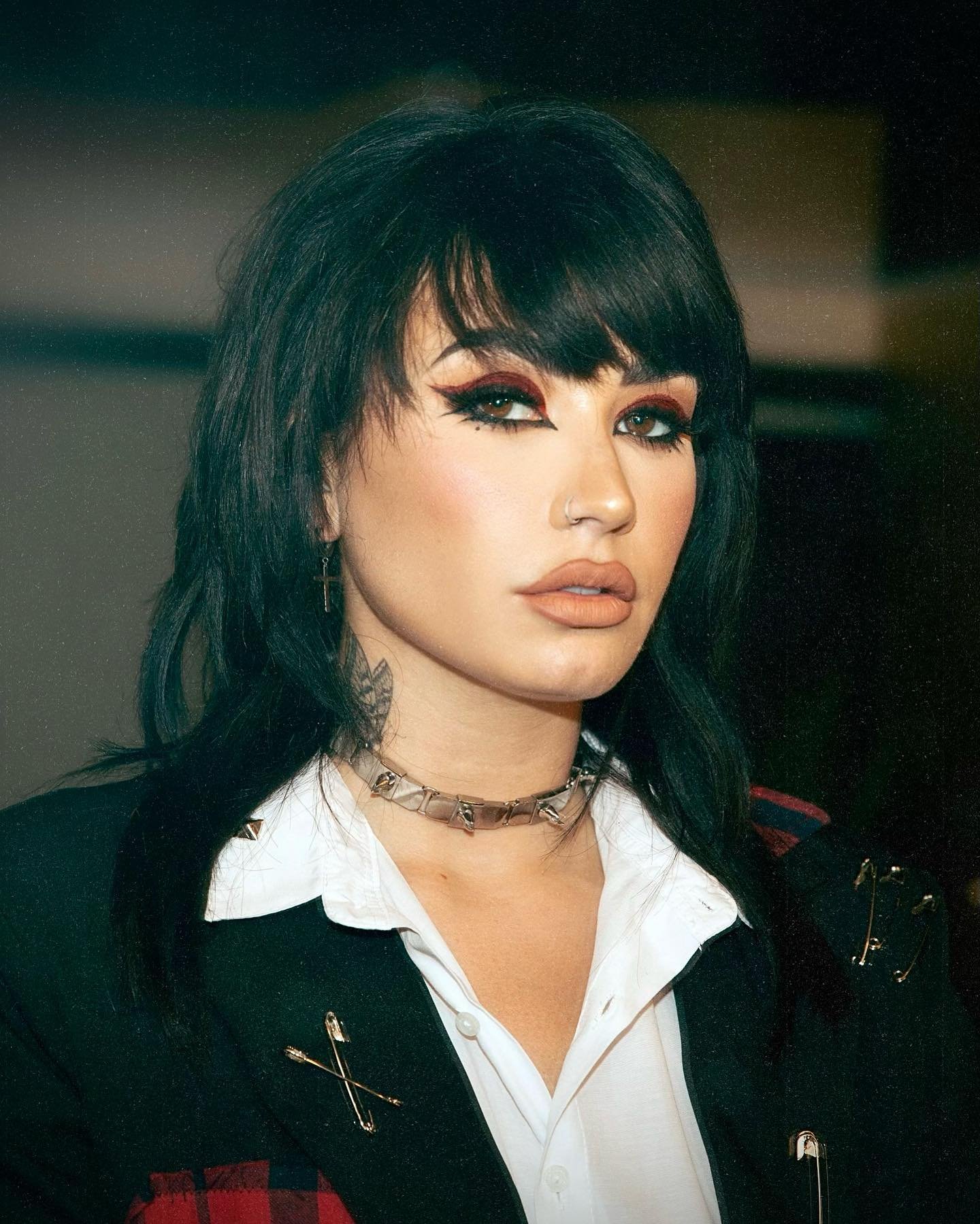 Portrait of a woman with dark hair, bold winged eyeliner, and a black blazer. She wears a metal choker necklace and posed expression.