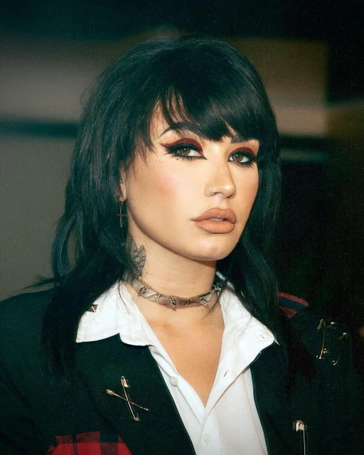 Portrait of a woman with dark hair, bold winged eyeliner, and a black blazer. She wears a metal choker necklace and posed expression.