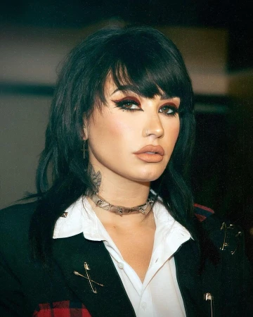 Portrait of a woman with dark hair, bold winged eyeliner, and a black blazer. She wears a metal choker necklace and posed expression.