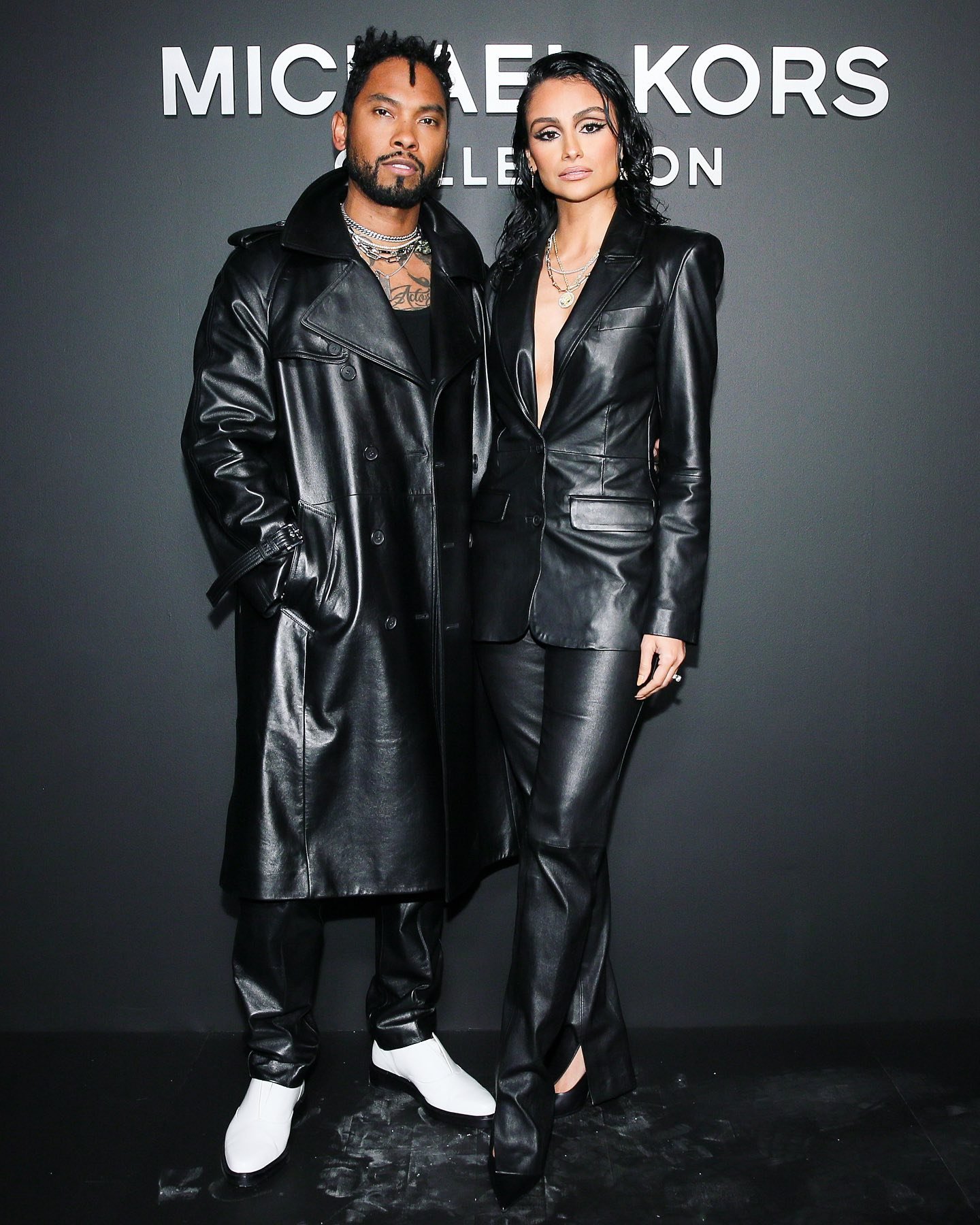 Two models pose against a dark background wearing leather outfits. The male wears a trench coat, the female wears a blazer.