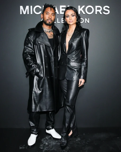 Two models pose against a dark background wearing leather outfits. The male wears a trench coat, the female wears a blazer.