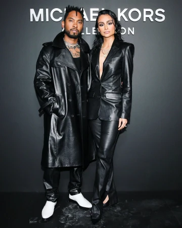Two models pose against a dark background wearing leather outfits. The male wears a trench coat, the female wears a blazer.