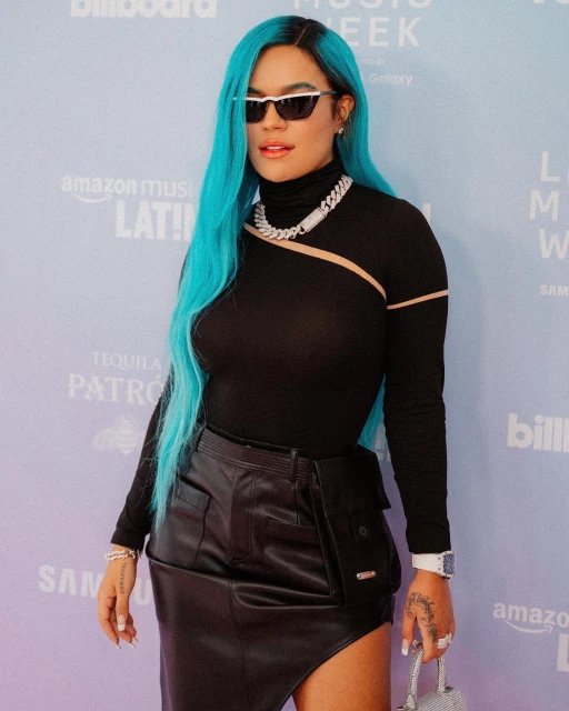 Karol G poses in a black top and skirt with turquoise hair, sunglasses, and jewelry against a Billboard Latin Music Week backdrop.