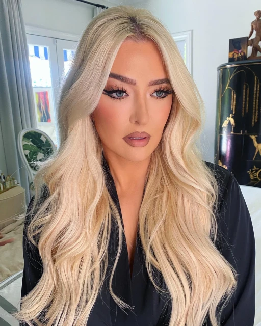 Portrait of an adult female with long blonde hair, wearing a dark robe. She has makeup on and is staring directly at the camera.
